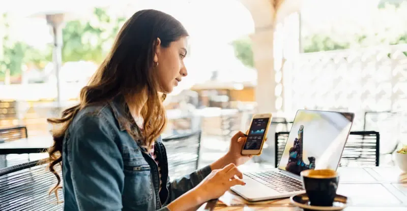 A woman sits at a cafe table, checking her phone while working on a laptop with a cup of coffee nearby